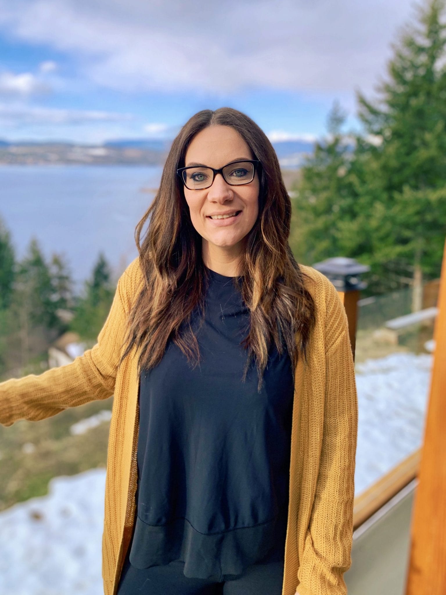 Emily Toews smiles directly at the camera, with pine trees and a lake in the background.
