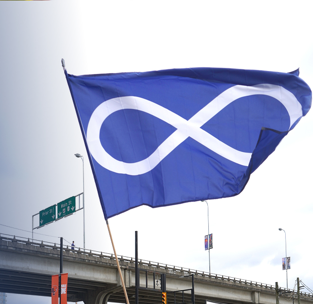 A man waving the blue Métis infinity sign flag outside in a parking lot.