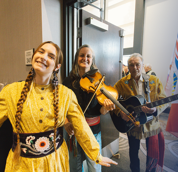 A woman is smiling and wearing traditional Métis clothing with a large black belt with a white beaded flower. Another woman is smiling, holding a fiddle, next to a man holding an acoustic guitar.