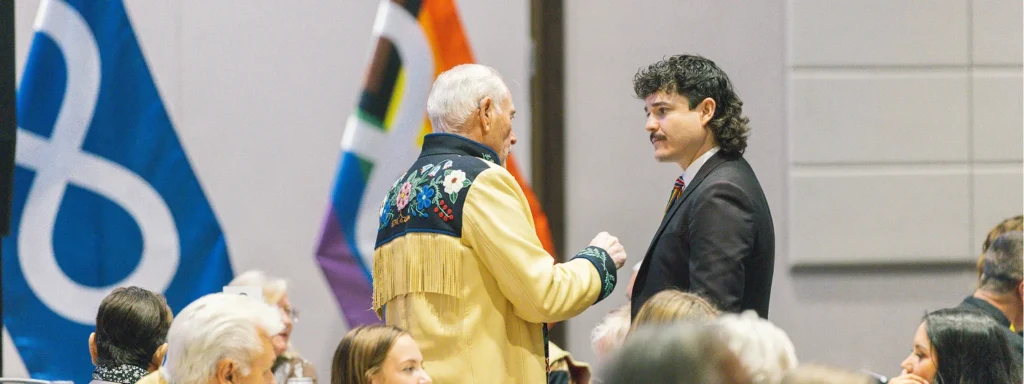 Two men are talking and standing behind other people sitting in a banquet hall.