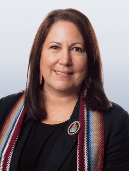 Carmen Carriere is smiling in a black sweater with a rainbow-coloured Métis sash and a beaded flower pin on her lapel against a white background.