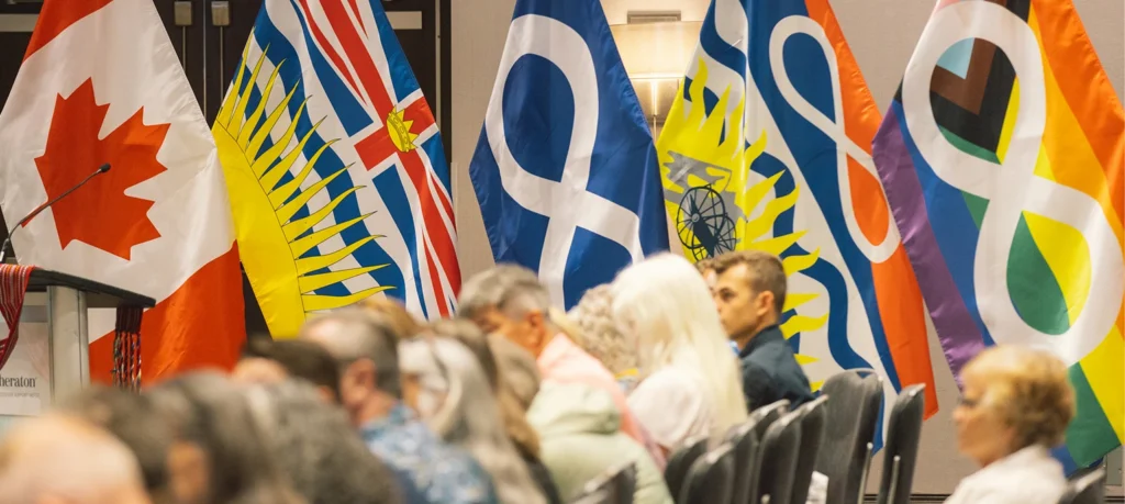 A group of people are sitting in rows of chairs at an event in front of a row of flags. The flags displayed are the Canadian flag, the flag of British Columbia, the Métis flag, a combined British Columbia and Métis flag, and a Pride Métis flag.