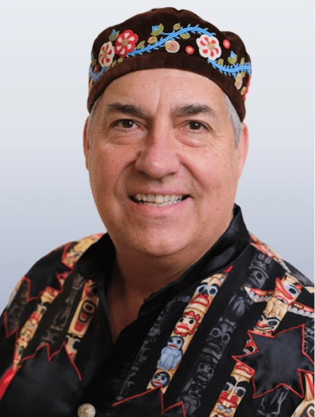 Louis De Jaeger is smiling in a black shirt with embroidered totem poles and is also wearing a black hat with beaded flowers on a white background.